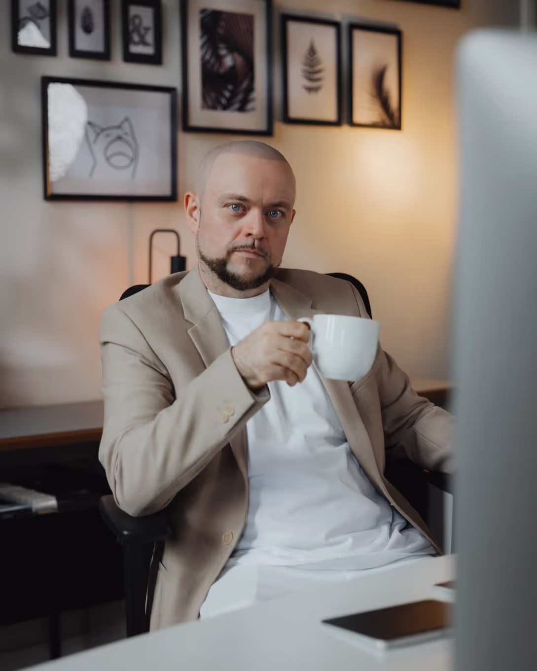 Ein Mann mit Glatze und kurzem Bart sitzt an einem Schreibtisch und hält eine große weiße Tasse in der Hand. Er trägt einen beigen Blazer über einem weißen T-Shirt. Im Hintergrund sind an der Wand mehrere gerahmte Bilder mit minimalistischen Designs und Pflanzenmotiven zu sehen. Der Mann schaut direkt in die Kamera und sitzt in einem modernen Büro mit einem Computerbildschirm vor sich.