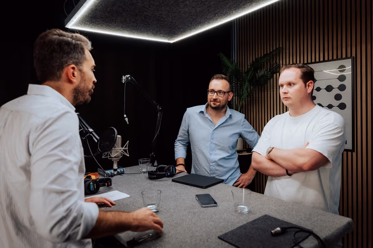 Drei Männer stehen in einem professionell eingerichteten Aufnahmeraum und unterhalten sich. Der Mann links trägt ein weißes Hemd und spricht zu den anderen beiden Männern, die ihm gegenüberstehen. Der Mann in der Mitte trägt ein hellblaues Hemd und eine Brille, während der Mann rechts ein weißes T-Shirt trägt. Auf dem Tisch vor ihnen stehen Mikrofone, Kopfhörer und Gläser mit Wasser. Der Raum hat eine moderne Akustikwand und eine Pflanze im Hintergrund. Eine helle Deckenbeleuchtung sorgt für eine gut ausgeleuchtete Szene.