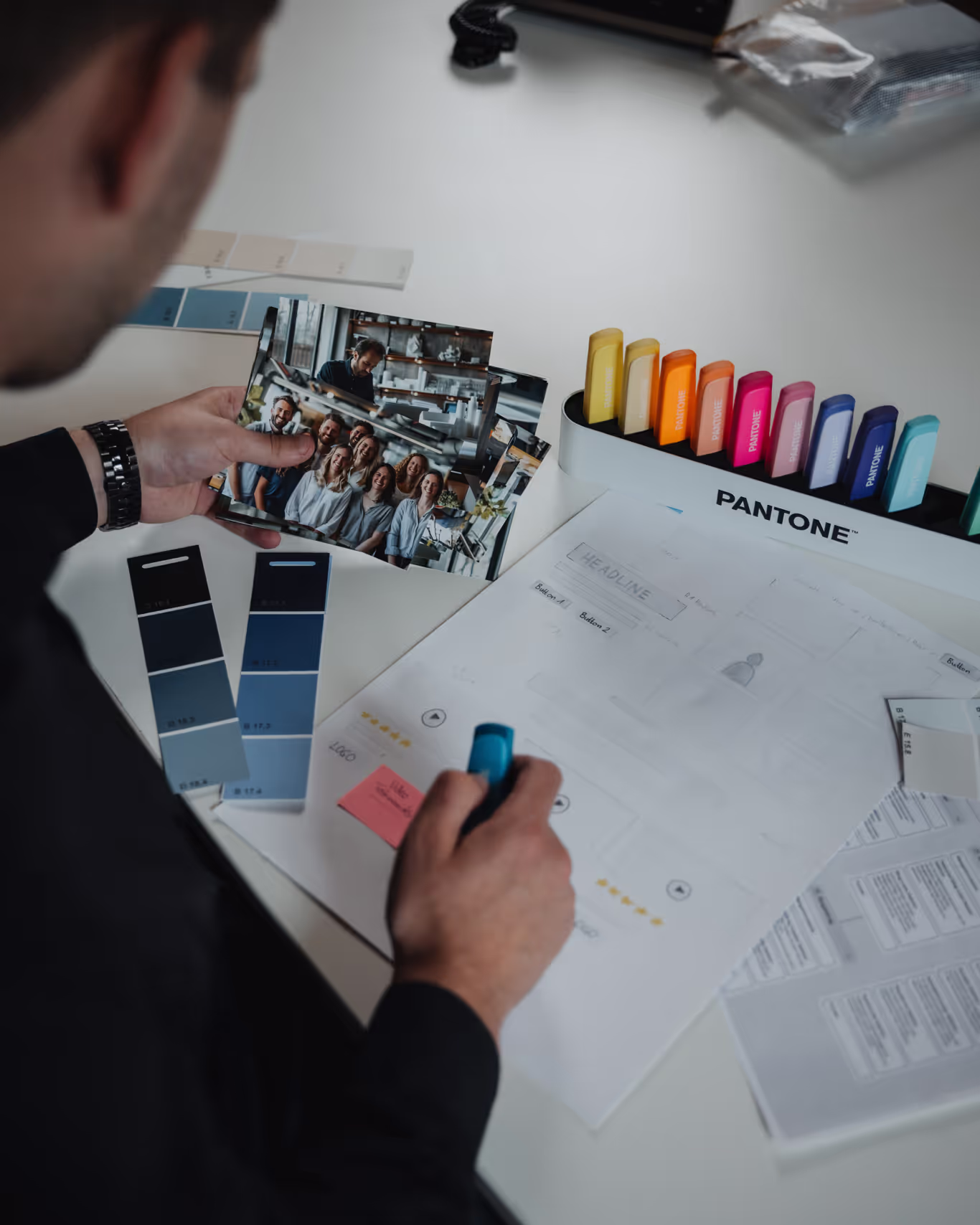 Person hält Fotos und umgeben von Pantone-Markern, Farbmusterkarten und Notizen auf einem weißen Tisch.
