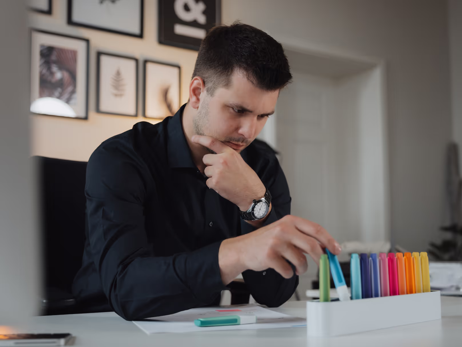Mann in schwarzem Hemd, der nachdenklich bunte Markerstifte aus einem Behälter auf einem Schreibtisch auswählt.