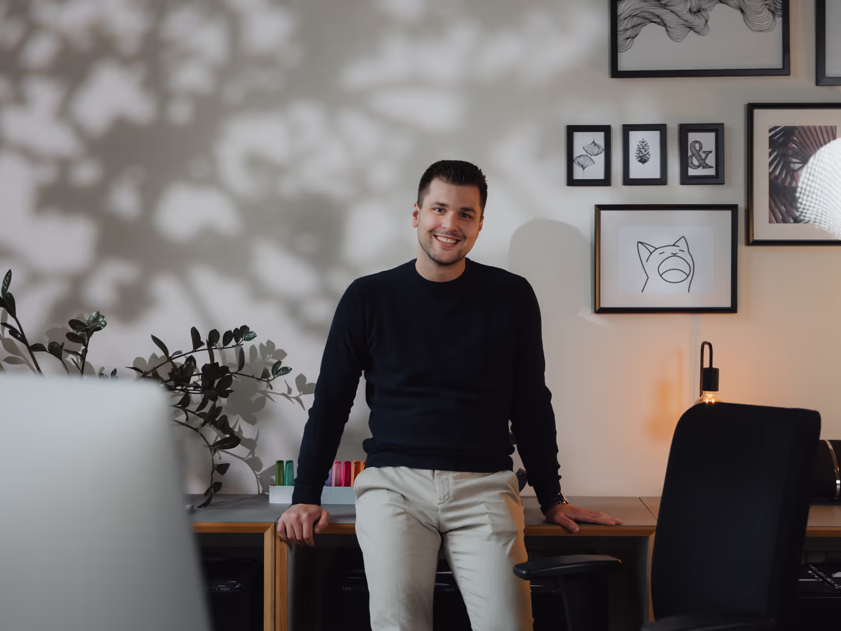 Junger Mann in schwarzem Pullover und beiger Hose, der auf einem Schreibtisch sitzt und lächelt, umgeben von dekorativen Wandbildern und Pflanzen.