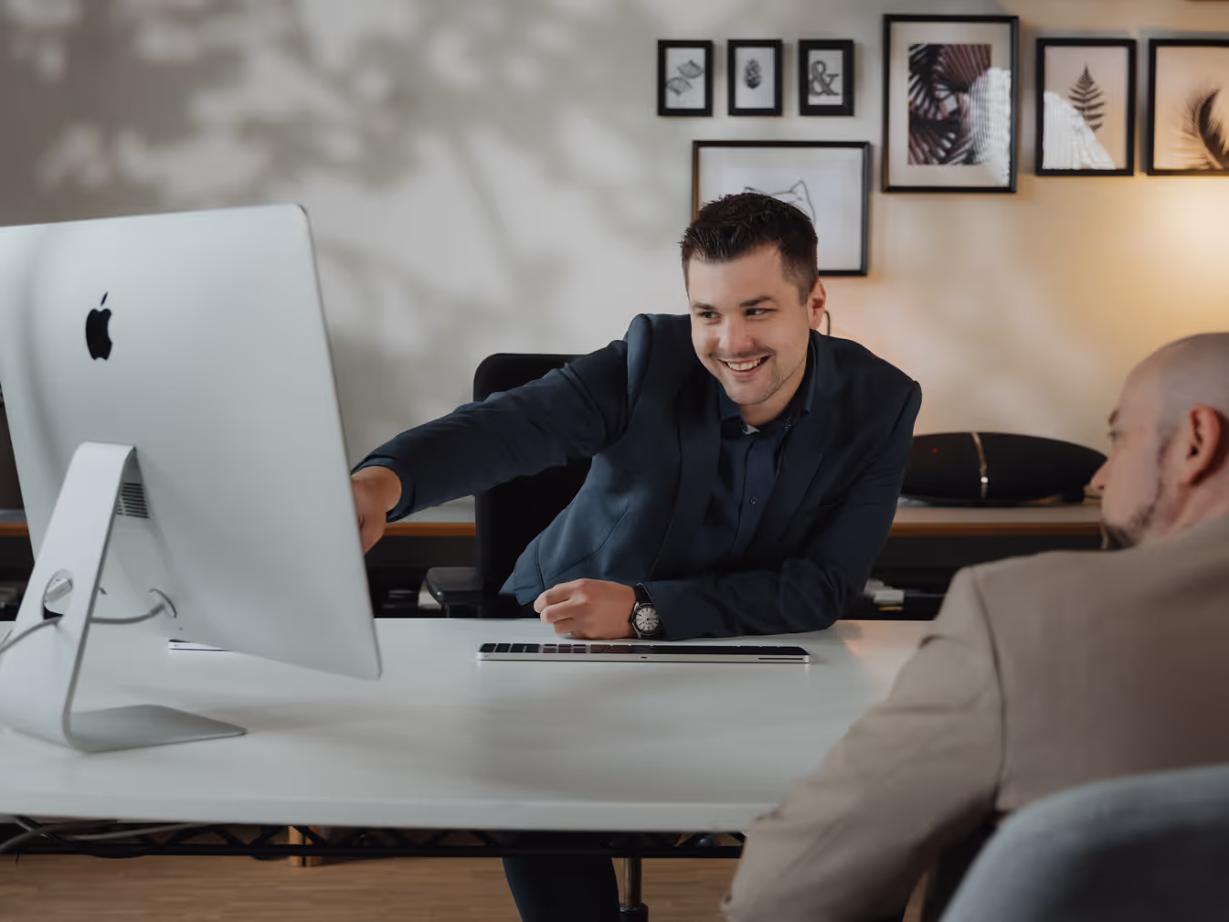 Junger Mann im Anzug zeigt lächelnd auf einen iMac-Bildschirm, während er mit einem anderen Mann in einem Büro spricht.