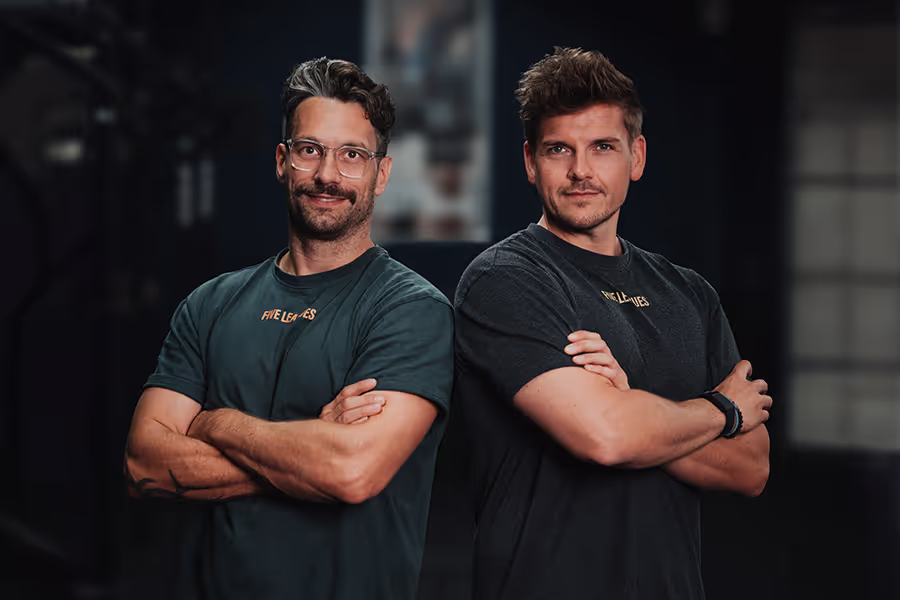 Zwei Männer stehen nebeneinander mit verschränkten Armen und blicken selbstbewusst in die Kamera. Beide tragen dunkle T-Shirts mit dem Logo “Five Leaves”. Der Hintergrund ist unscharf, wodurch der Fokus klar auf den beiden Personen liegt, die in einer entspannten, aber kraftvollen Pose posieren.