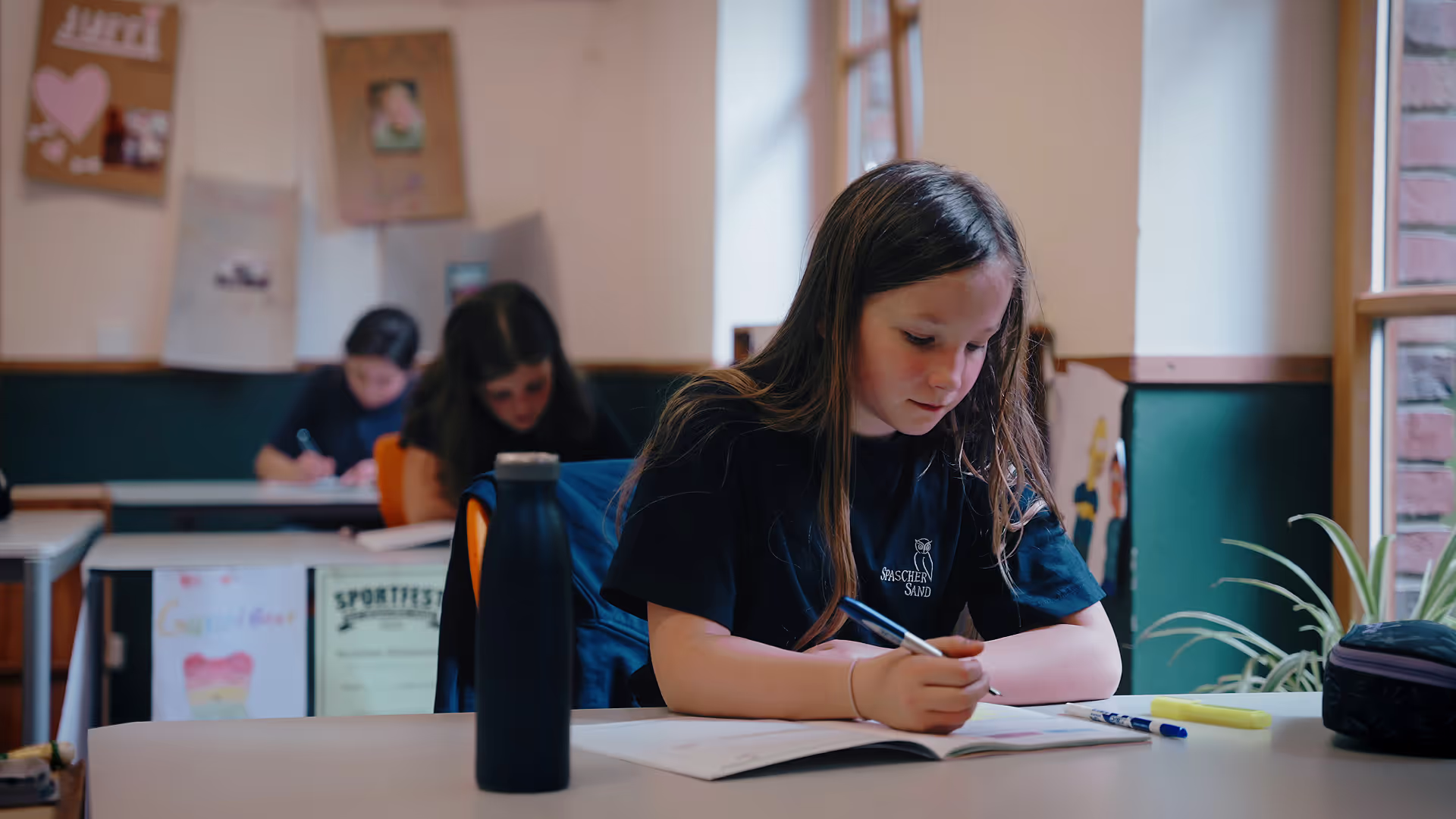 Ein junges Mädchen sitzt in einem Klassenzimmer an einem Tisch und konzentriert sich auf das Schreiben in ein Heft. Im Hintergrund sind zwei weitere Kinder zu sehen, die ebenfalls an ihren Tischen arbeiten. Das Klassenzimmer ist hell und freundlich eingerichtet, mit Pflanzen und Kunstwerken an den Wänden. Auf dem Tisch des Mädchens liegen Schreibutensilien und eine Trinkflasche.