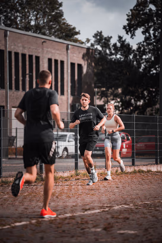 Drei Personen joggen auf einer Laufbahn im Freien. Im Vordergrund ist ein Mann von hinten zu sehen, der auf zwei weitere Läufer zuläuft. Die beiden anderen Läufer, ein Mann und eine Frau, befinden sich in der Mitte des Bildes und joggen in dieselbe Richtung. Sie tragen sportliche Kleidung und sind konzentriert. Im Hintergrund sind ein Backsteingebäude und parkende Autos zu sehen. Die Szene wirkt dynamisch und sportlich, mit einem leicht bewölkten Himmel im Hintergrund.