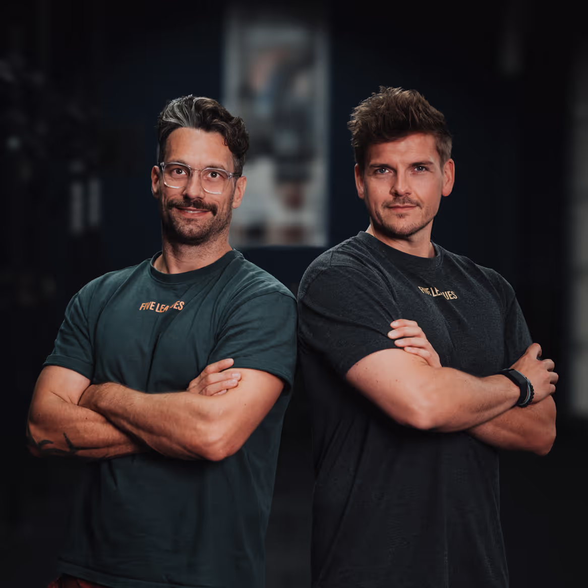 Zwei Männer stehen nebeneinander mit verschränkten Armen und blicken selbstbewusst in die Kamera. Beide tragen T-Shirts mit dem Logo “Five Leagues”. Der linke Mann trägt eine Brille und hat kurzes, welliges Haar, während der rechte Mann ebenfalls kurzes Haar hat und eine Armbanduhr trägt. Der Hintergrund ist dunkel und unscharf, wodurch der Fokus klar auf den beiden Personen liegt. Sie posieren in einer entspannten, aber entschlossenen Haltung.