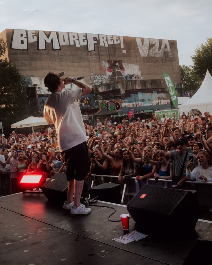 Ein junger Mann mit Baseballkappe und tätowierten Armen steht auf einer Bühne und hält ein Mikrofon in der Hand. Er trägt ein T-Shirt und blickt konzentriert nach unten, als würde er sich auf seine Performance vorbereiten. Im Hintergrund sind verschwommene Bühnenelemente und Lichteffekte zu erkennen, was auf eine Konzert- oder Eventkulisse hindeutet. Die Atmosphäre wirkt energiegeladen und fokussiert.