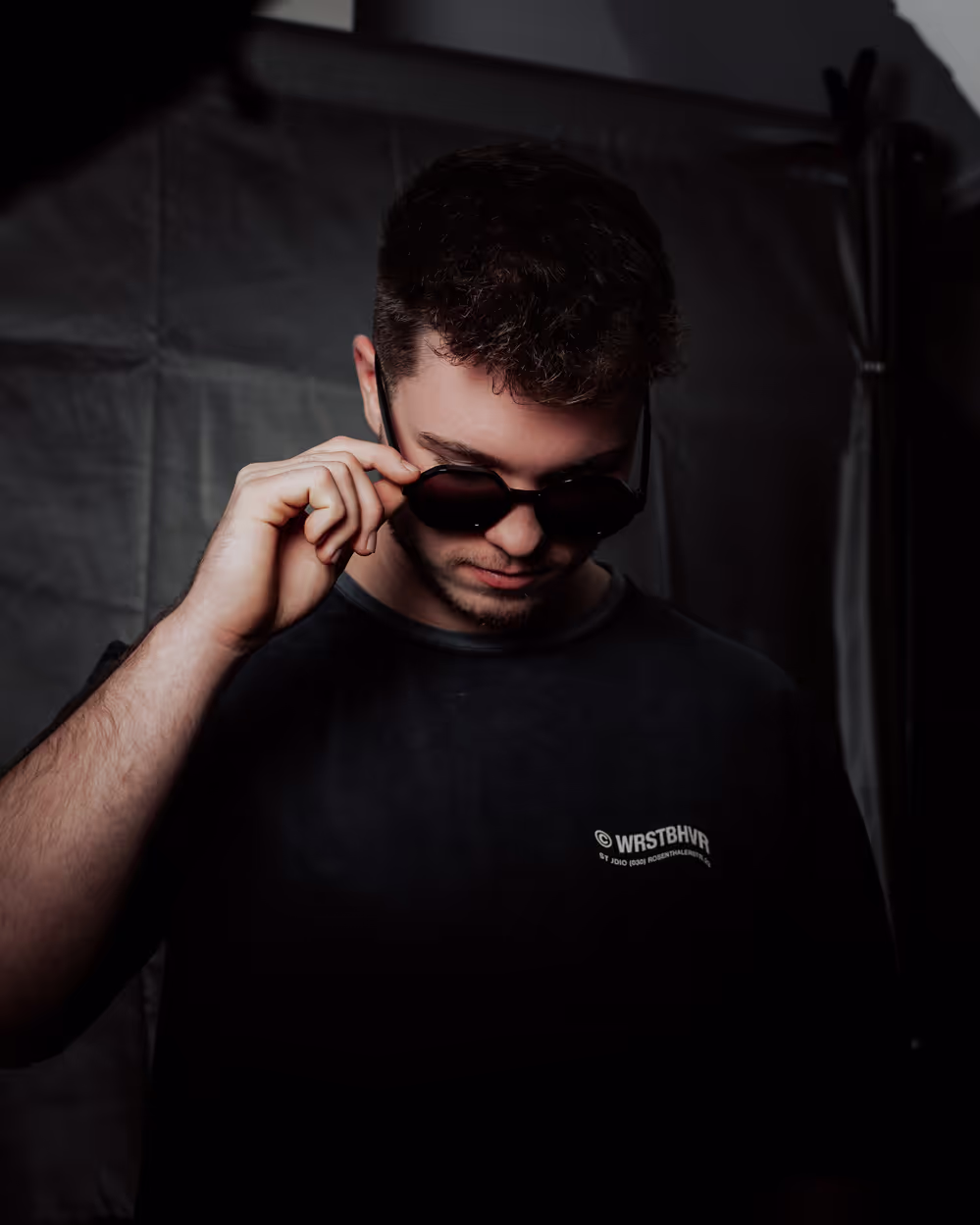 Ein Mann mit kurzem Haar und Bart posiert vor einem dunklen Hintergrund. Er trägt ein Hemd und schaut selbstbewusst in die Kamera. Die Szene ist schlicht und der Fokus liegt auf seinem Gesicht, wodurch ein professioneller und fokussierter Eindruck entsteht.