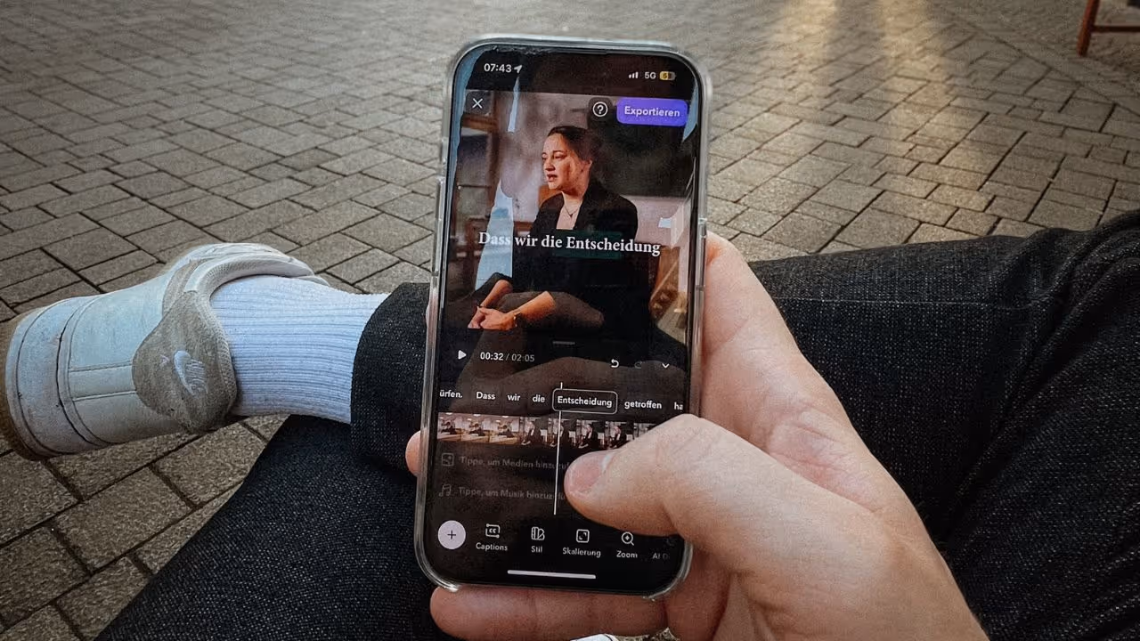Das Bild zeigt eine Person, die ein Smartphone in der Hand hält und offenbar gerade ein Video schneidet oder bearbeitet. Die Szene wirkt entspannt – die Person trägt weiße Socken, weiße Slipper und dunkle Jeans, sitzt vermutlich draußen auf einem gepflasterten Platz. Auf dem Handybildschirm ist ein Video einer Frau zu sehen, darüber der Text: „Dass wir die Entscheidung…“. Oben rechts befindet sich ein Button mit der Aufschrift „Exportieren“, was darauf hinweist, dass die Bearbeitung fast abgeschlossen ist.