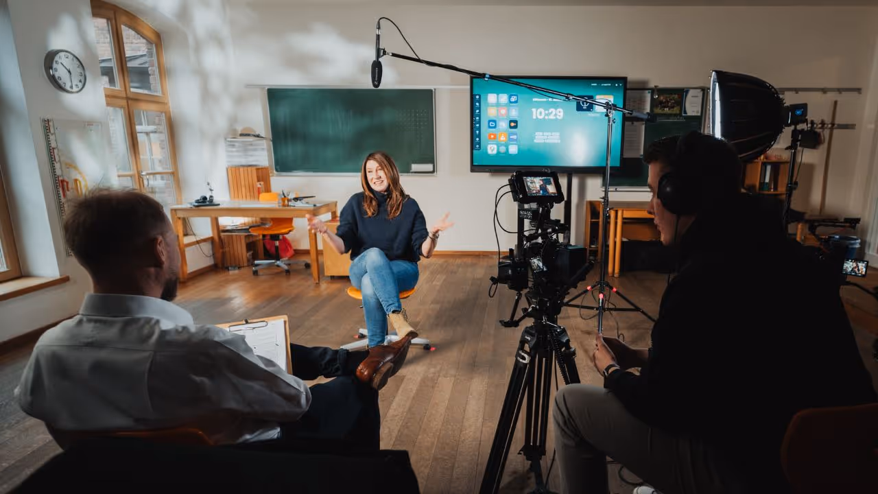 In einem hellen Klassenraum mit Holzboden und großen Fenstern wird ein Interview gefilmt. Eine Frau sitzt locker auf einem orangefarbenen Stuhl und spricht gestikulierend in die Kamera. Vor ihr sitzt ein Mann mit Klemmbrett, rechts im Bild filmt ein Kameramann mit professionellem Equipment. Über der Frau hängt ein Tonangel-Mikrofon, im Hintergrund ist ein interaktives Display mit Uhrzeit (10:29) sowie eine grüne Tafel zu sehen. Die Szene wirkt professionell und authentisch – vermutlich handelt es sich um die Produktion eines Imagefilms für eine Schule oder Bildungseinrichtung.