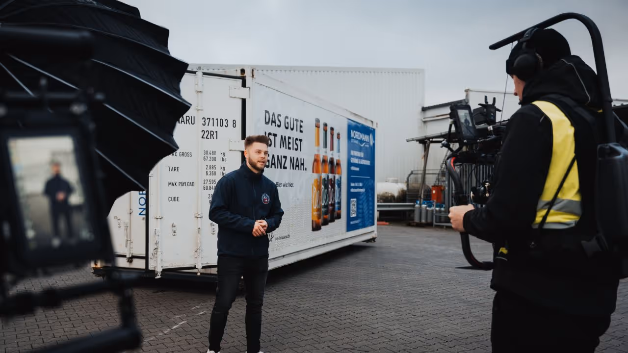 Ein junger Mann steht vor einem großen Container mit Bierwerbung und spricht direkt in die Kamera. Er wird von einem professionellen Kamerateam mit Gimbal, Licht und Ton-Equipment gefilmt. Im Hintergrund ist ein Industriegelände mit Lagerflächen zu sehen. Die Szene wirkt wie ein authentischer Werbefilmdreh im Business-Umfeld.