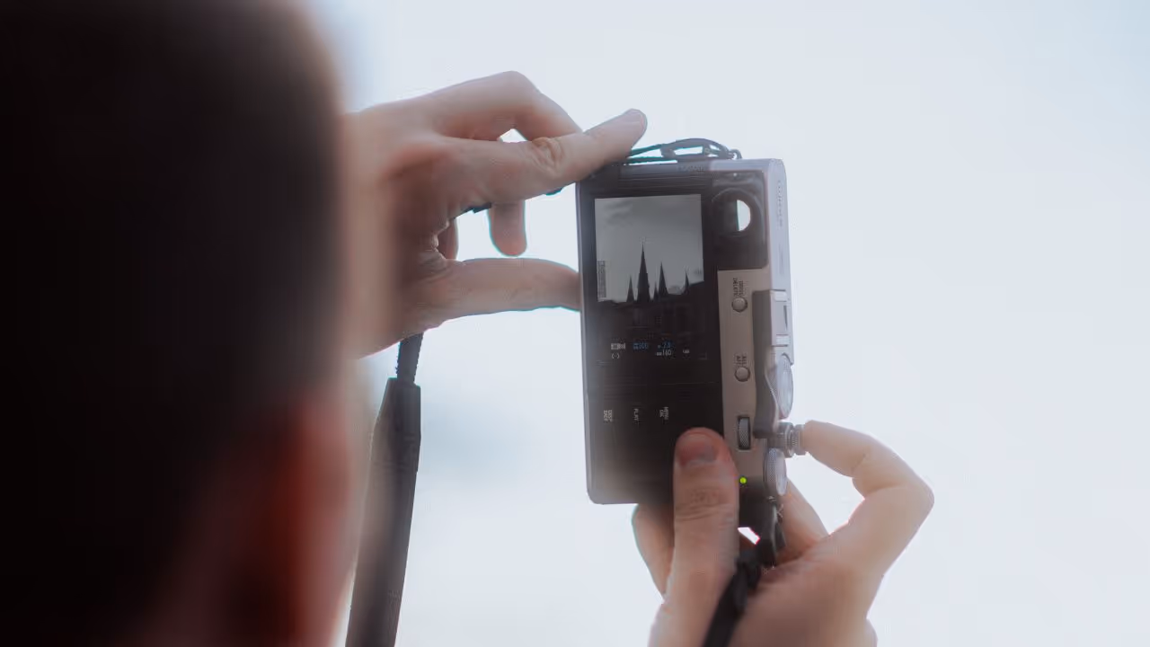 Eine Person hält eine kompakte Digitalkamera in den Händen und fotografiert oder filmt eine markante Skyline mit spitzen Gebäudespitzen – möglicherweise eine Kathedrale. Die Kamera zeigt das Motiv im Schwarzweißmodus auf dem Display, während der Hintergrund stark überbelichtet und weiß erscheint. Die Szene wirkt minimalistisch und künstlerisch.
